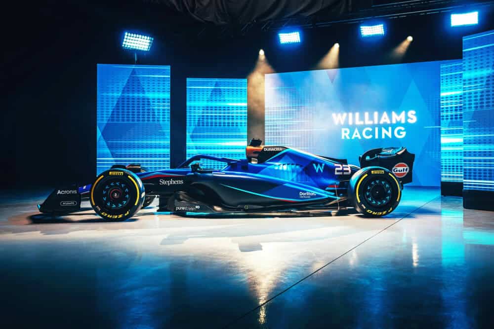La imagen muestra un coche de Fórmula 1 del equipo Williams Racing, exhibido en un entorno bien iluminado con una temática de iluminación azul. El coche presenta un diseño elegante con una combinación de colores predominantemente azul y negro, adornado con varios logotipos de patrocinadores como Acronis, Stephens, Duracell, Dorilton y Gulf. El número "23" es visible en el coche, y los neumáticos están marcados con la marca Pirelli. En el fondo, se puede ver el texto "WILLIAMS RACING" iluminado en blanco contra un fondo azul con patrones geométricos. Esta imagen es interesante y relevante ya que muestra el diseño y los patrocinadores del coche de Fórmula 1 del equipo Williams Racing, probablemente para una nueva temporada o un evento de presentación.