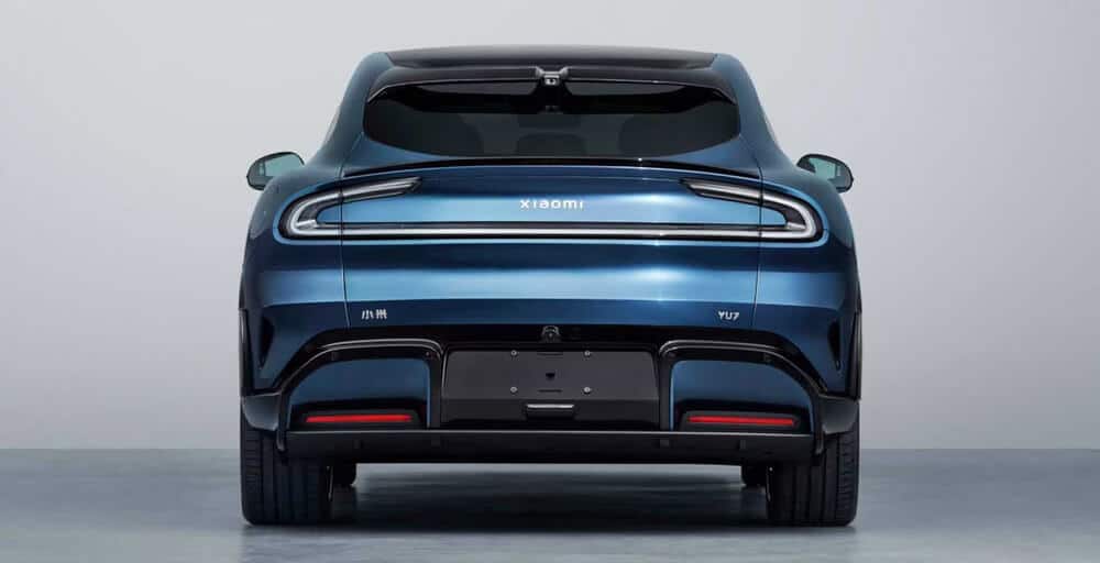 La imagen muestra la vista trasera de un coche azul Xiaomi YU7. El coche presenta elementos de diseño modernos y elegantes, incluyendo una tira continua de luces LED traseras, un limpiaparabrisas trasero y salidas de escape dobles. El logotipo de Xiaomi se muestra de manera prominente en el centro de la parte trasera, justo encima de la tira de luces traseras. El diseño del coche es interesante y relevante ya que representa la entrada de Xiaomi en el mercado automotriz, mostrando su expansión más allá de la electrónica de consumo hacia los vehículos eléctricos.