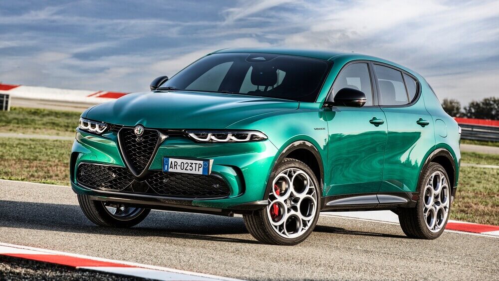 La imagen muestra un Alfa Romeo Tonale verde, un SUV compacto de lujo, estacionado en una pista de carreras. El coche presenta un diseño elegante y moderno con elementos distintivos de Alfa Romeo, como la parrilla en forma de V y las llantas deportivas de aleación. La matrícula dice "AR-023TP". El entorno sugiere un enfoque en el rendimiento, con bordillos rojos y blancos y un área abierta con césped en el fondo. Esta imagen es interesante y relevante porque destaca el diseño y el entorno orientado al rendimiento del vehículo, subrayando su atractivo deportivo y sus capacidades de conducción.