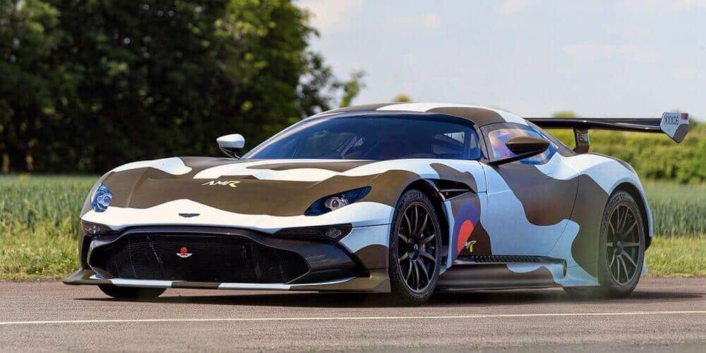 La imagen muestra un Aston Martin Vulcan con matrícula para calle. El coche presenta un distintivo diseño de camuflaje en tonos de marrón, blanco y azul, con líneas aerodinámicas elegantes, un gran alerón trasero y entradas de aire frontales prominentes, lo que indica su diseño para velocidad y rendimiento. El coche está estacionado en una carretera con vegetación en el fondo, sugiriendo que está al aire libre, posiblemente en una pista o en un lugar pintoresco. La matrícula dice "XXX06." Esta imagen es interesante y relevante porque muestra un coche deportivo diseñado para alto rendimiento, destacando tanto su estética como sus capacidades técnicas.