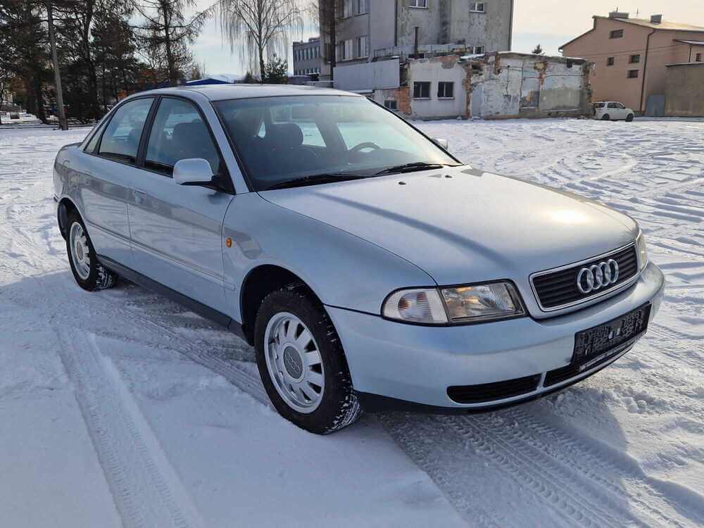 La imagen muestra un Audi A4 B5 plateado estacionado sobre una superficie nevada. El coche está posicionado en un ángulo que muestra el frente y el lado del conductor. El logotipo de Audi se muestra de manera prominente en la parrilla frontal. En el fondo, se pueden ver algunos edificios y árboles, lo que indica un entorno urbano o suburbano. La nieve en el suelo y en los neumáticos del coche sugiere condiciones invernales. Esta imagen es interesante ya que destaca el diseño y la condición del coche en un entorno invernal, lo cual podría ser relevante para mostrar el rendimiento o la durabilidad del vehículo en climas fríos.