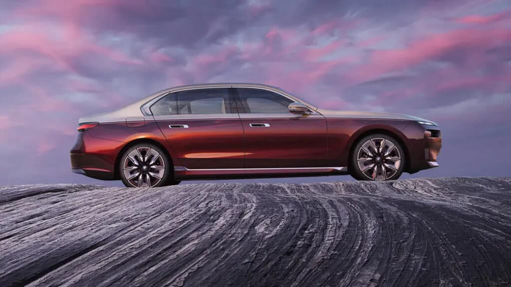 "Vista lateral de un sedán de lujo, específicamente el BMW i7, posicionado en un terreno rocoso con un cielo dramático de fondo. El coche tiene un diseño elegante y moderno con un esquema de colores en dos tonos, presentando una carrocería metálica roja y un techo plateado. Las ruedas son grandes y estilizadas, complementando la estética general del vehículo. El cielo de fondo tiene una mezcla de tonos rosados y púrpuras, añadiendo atractivo visual a la imagen. Esta imagen es interesante y relevante ya que muestra el diseño y la elegancia del BMW i7, destacando su presencia en un entorno pintoresco.