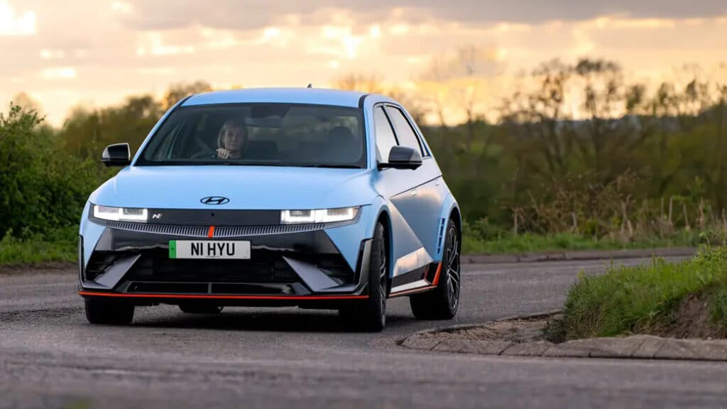 Un Hyundai Ioniq 5 N, un vehículo eléctrico de alto rendimiento, conduciendo por una carretera con un fondo escénico de árboles y un cielo al atardecer. El coche está pintado de color azul claro con acentos negros y naranjas, y tiene un diseño frontal distintivo con faros LED y una apariencia deportiva. La matrícula dice 'N1 HYU'. Esta imagen es interesante y relevante porque destaca el diseño y las características del Ioniq 5 N, mostrando su estética deportiva y moderna en un entorno natural.