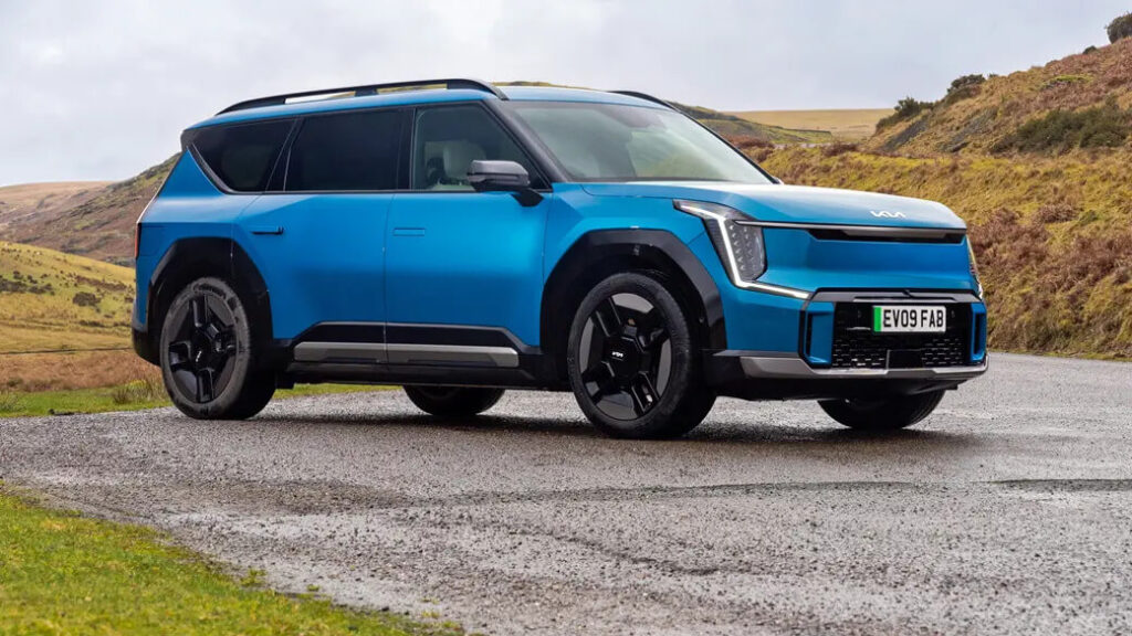Un KIA EV9 azul, un SUV eléctrico, estacionado en una carretera pavimentada con un paisaje escénico y montañoso al fondo. El vehículo tiene un diseño moderno y robusto con faros LED distintivos y ruedas de aleación negras. La matrícula dice 'EV09 FAB'. Esta imagen es interesante y relevante ya que muestra el diseño y el atractivo estético del KIA EV9, destacando su presencia en un entorno natural, lo que puede atraer a posibles compradores interesados en SUVs eléctricos.