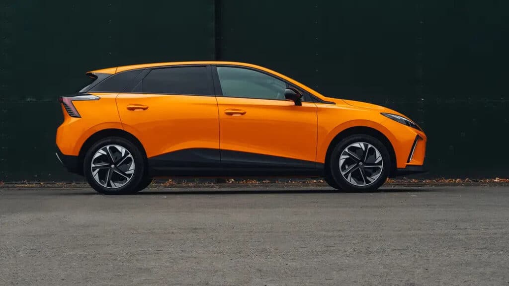 Vista lateral de un MG4 naranja. El coche tiene un diseño elegante y moderno con líneas afiladas y una apariencia deportiva. Las ruedas tienen un patrón único y el coche presenta acentos negros a lo largo de la parte inferior de la carrocería y alrededor de las ventanas. El fondo es una pared oscura y lisa, lo que hace que el color brillante del coche destaque.