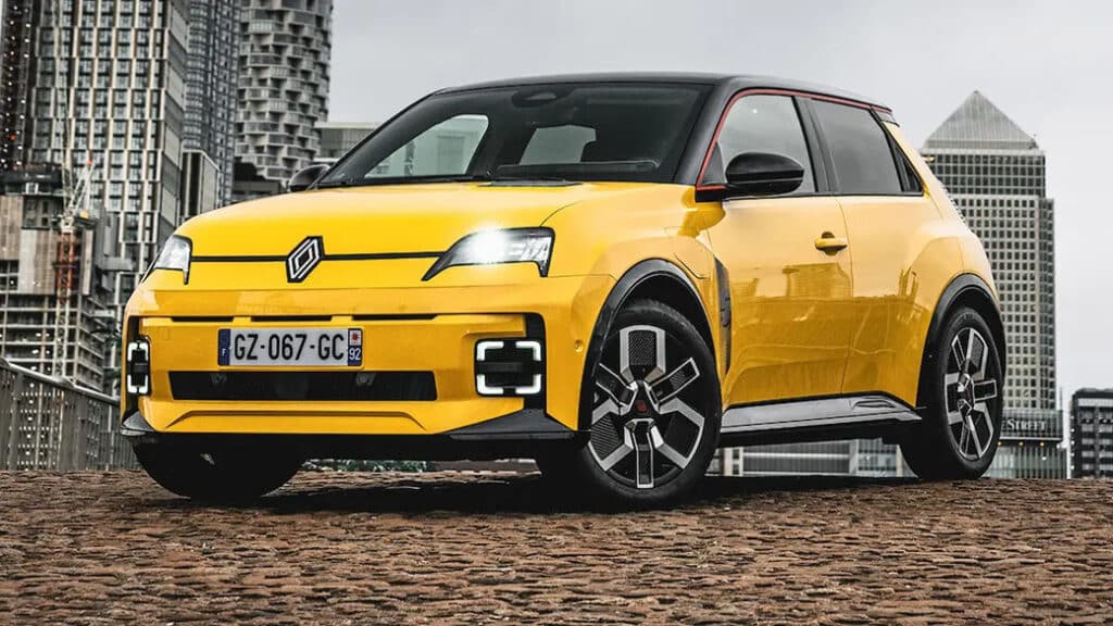 "Un Renault 5 eléctrico amarillo estacionado sobre una superficie de adoquines con un paisaje urbano de fondo. El coche tiene un diseño moderno y deportivo con faros LED distintivos y un techo negro. La matrícula del coche dice 'GZ-067-GC'. El diseño del coche y el entorno urbano lo hacen visualmente atractivo y relevante para mostrar el diseño automotriz moderno en un entorno urbano.