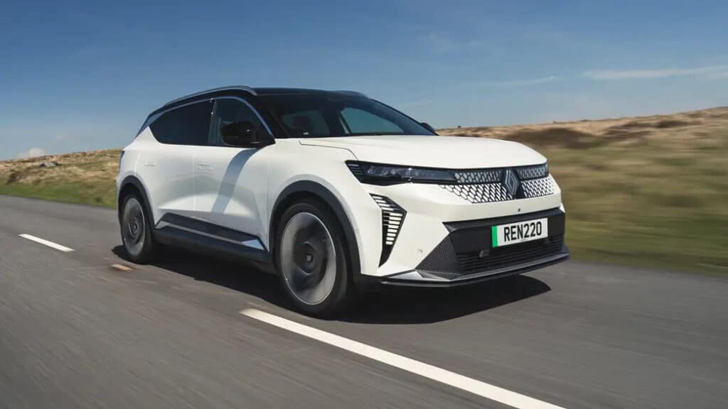 "Un Renault Scenic blanco conduciendo por una carretera. El vehículo tiene un diseño moderno y elegante con faros delanteros distintivos y una parrilla única. La matrícula dice 'REN220'. El fondo muestra un paisaje abierto con un cielo azul claro, sugiriendo que el coche está siendo conducido en un entorno rural o campestre. Esta imagen es interesante ya que destaca el diseño y la estética del Renault Scenic, mostrando sus características en un escenario dinámico de conducción.