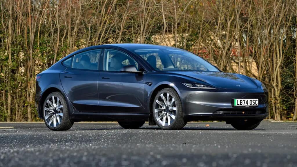 "Un Tesla Model 3 gris oscuro estacionado en una carretera con un fondo de árboles sin hojas. El coche es un vehículo eléctrico moderno y elegante. La matrícula del coche dice 'LB74 OSC'. Esta imagen es interesante y relevante ya que muestra el diseño y la estética del Tesla Model 3, un vehículo eléctrico popular conocido por su tecnología avanzada y beneficios ambientales.
