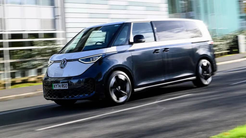 "Un Volkswagen ID. Buzz, una furgoneta eléctrica, conduciendo por una carretera. El vehículo destaca por su diseño moderno, con líneas elegantes, un esquema de colores en dos tonos y luces delanteras y traseras distintivas. La matrícula dice 'KY74 RGO'. Esta imagen es interesante porque resalta el resurgimiento de la icónica furgoneta Volkswagen en una forma eléctrica, mostrando avances en tecnología y diseño automotriz.