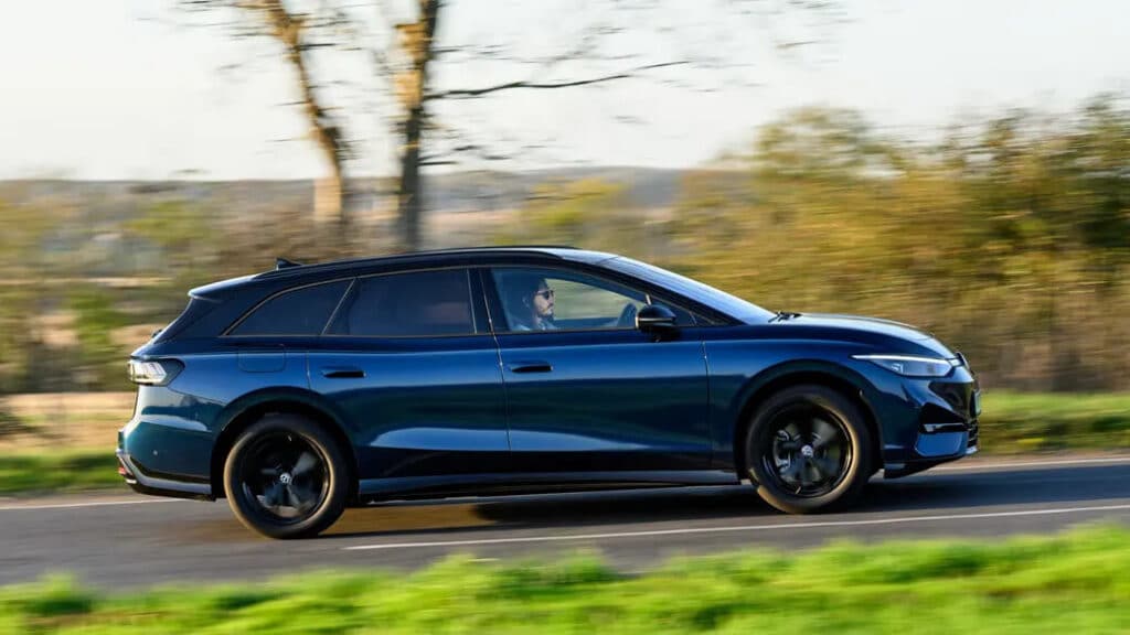 Vista lateral de un Volkswagen ID.7 Tourer azul conduciendo por una carretera. El coche está en movimiento, y el fondo está ligeramente borroso, lo que indica velocidad. El vehículo tiene un diseño elegante y moderno con ruedas de aleación negras y ventanas tintadas. El conductor es visible a través de la ventana. Esta imagen es interesante y relevante ya que muestra el diseño y la funcionalidad de un vehículo eléctrico, destacando su potencial como opción de transporte sostenible.