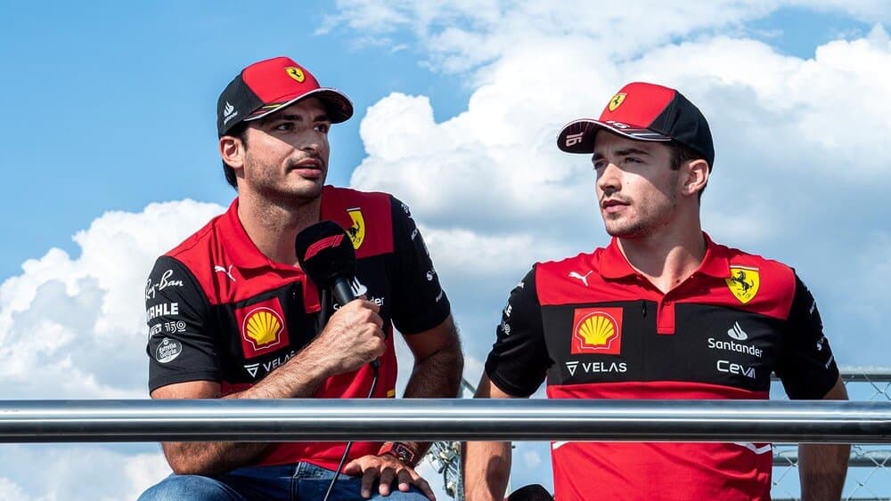 La imagen muestra a Carlos Sainz Jr. y Charles Leclerc, ambos pilotos de Fórmula 1 del equipo Ferrari, vistiendo uniformes rojos con logotipos de patrocinadores como Shell, Velas, Santander y Ceva. Sainz está sosteniendo un micrófono, lo que sugiere que podrían estar dando una entrevista o hablando en un evento. El fondo muestra un cielo azul con nubes blancas, indicando que están al aire libre. Esta imagen es interesante y relevante ya que captura a dos destacados pilotos de Ferrari en un contexto de interacción con los medios o el público.