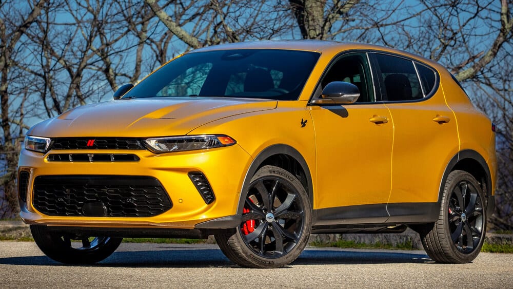 La imagen muestra un Dodge Hornet amarillo, un SUV compacto, estacionado al aire libre sobre una superficie pavimentada con árboles sin hojas en el fondo. El Dodge Hornet presenta un diseño elegante con una parrilla frontal prominente, llantas de aleación negras y pinzas de freno rojas. Esta imagen es interesante y relevante porque el Dodge Hornet es conocido por ser una versión más asequible del Alfa Romeo Tonale, compartiendo muchos elementos de diseño e ingeniería con él.