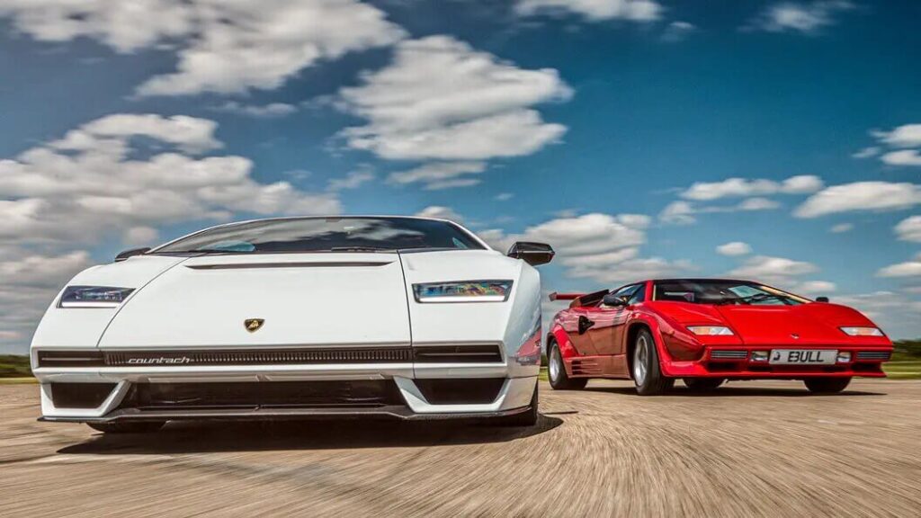 La imagen muestra dos coches Lamborghini Countach conduciendo por una carretera bajo un cielo parcialmente nublado. El coche en primer plano es blanco y tiene un diseño moderno con líneas afiladas y una apariencia elegante. El coche en el fondo es rojo y tiene un diseño clásico con un aspecto más angular y retro. La imagen es interesante porque yuxtapone las versiones moderna y clásica del icónico Lamborghini Countach, destacando la evolución del diseño del coche a lo largo del tiempo. La matrícula del coche rojo es "E3 ULL".