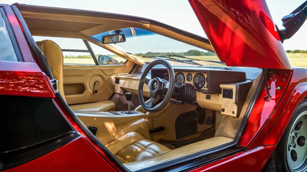 "Imagen del interior de un Lamborghini Countach clásico con exterior rojo. El coche tiene un interior de cuero color beige, incluyendo los asientos, el tablero y los paneles de las puertas. El volante está en el lado derecho, lo que indica que este coche está diseñado para un país donde se conduce por la derecha. El tablero presenta varios indicadores analógicos, incluyendo un velocímetro y un tacómetro. La puerta del coche está abierta, revelando el diseño detallado y lujoso del interior.