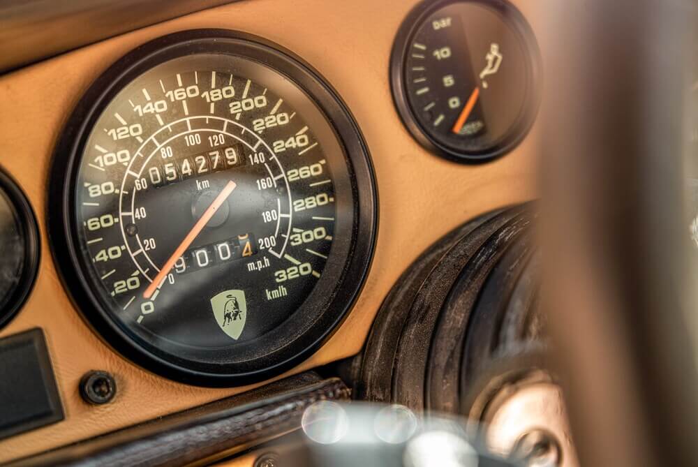La imagen muestra el tablero de un Lamborghini Countach clásico. El velocímetro tiene un rango de 0 a 320 km/h e incluye una escala más pequeña para mph, que va de 0 a 200 mph. El odómetro muestra una lectura de 054279 km. A la derecha del velocímetro, hay otro indicador que mide la presión en bar, con un rango de 0 a 10 bar. Esta imagen es interesante porque destaca la instrumentación de un coche clásico, mostrando su diseño y las capacidades de alta velocidad indicadas por el velocímetro.
