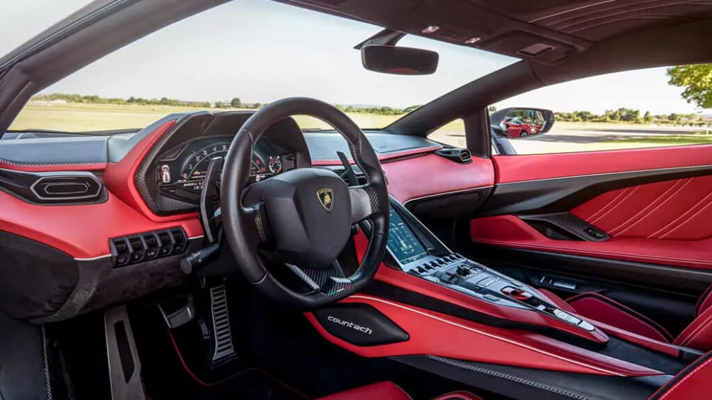 "Imagen del interior de un Lamborghini Countach LPI 800-4 nuevo con exterior rojo. El coche tiene un interior de cuero color beige, incluyendo los asientos, el tablero y los paneles de las puertas. El volante está en el lado derecho, lo que indica que este coche está diseñado para un país donde se conduce por la derecha. El tablero presenta varios indicadores analógicos, incluyendo un velocímetro y un tacómetro. La puerta del coche está abierta, revelando el diseño detallado y lujoso del interior.