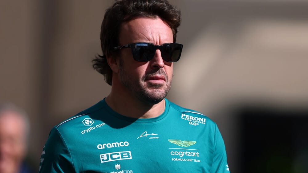 La imagen muestra a Fernando Alonso, piloto de Fórmula 1, con una camiseta verde que tiene varios logotipos y textos, incluyendo "crypto.com," "aramco," "JCB," "cognizant FORMULA ONE TEAM," "PERONI Nastro Azzurro 0.0%," y "Aston Martin." Fernando Alonso también lleva gafas de sol, lo que le da un aspecto muy jefe. Esta imagen es interesante y relevante porque está relacionada con el equipo de Fórmula 1 Aston Martin y el piloto Fernando Alonso, destacando su estilo y presencia en el mundo del automovilismo.