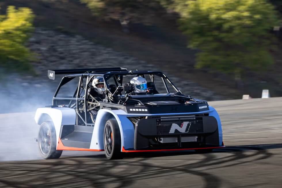 La imagen muestra un coche de carreras de alto rendimiento, específicamente el Hyundai RN24, derrapando en una pista. El coche tiene un diseño distintivo con una jaula antivuelco y características aerodinámicas, incluyendo un gran alerón trasero. El coche está marcado con la letra "N" en el alerón trasero y en el panel trasero. La pista tiene una curva y hay gradas vacías en el fondo, indicando un lugar de carreras. La escena se desarrolla durante el día con cielos despejados. Esta imagen es interesante porque captura un vehículo de alto rendimiento en un entorno de carreras, destacando los aspectos de ingeniería y diseño de los coches de carreras.