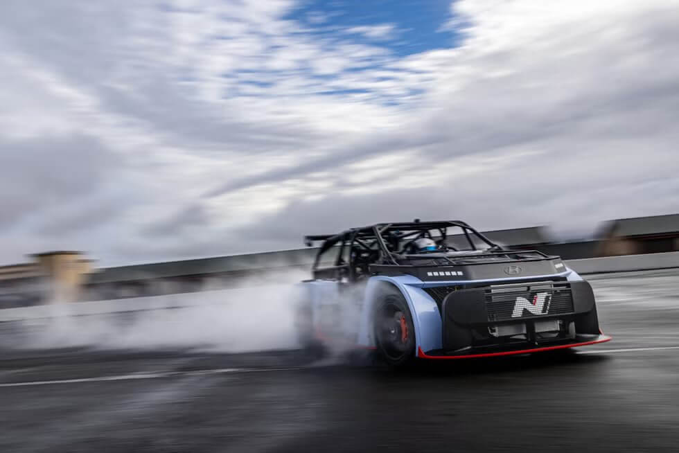 La imagen muestra un coche de carreras de alto rendimiento, específicamente el Hyundai RN22e, derrapando en una pista. El coche está emitiendo humo de sus neumáticos, lo que indica un movimiento a alta velocidad y una conducción agresiva. El coche tiene un diseño aerodinámico con una parrilla frontal prominente que presenta la letra "N". El fondo muestra un cielo nublado y algunas estructuras borrosas, sugiriendo que la escena tiene lugar en una instalación de carreras o pista. Esta imagen es interesante porque captura el movimiento dinámico y la potencia del coche de carreras, destacando sus capacidades y la habilidad requerida para controlarlo.