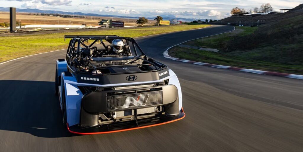La imagen muestra un coche de carreras de alto rendimiento, específicamente el Hyundai RN24, en una pista. El coche tiene un diseño distintivo con una jaula antivuelco y características aerodinámicas, incluyendo un gran alerón trasero. El coche está marcado con la letra "N" en el alerón trasero y en el panel trasero. La pista tiene una curva y hay gradas vacías en el fondo, indicando un lugar de carreras. La escena se desarrolla durante el día con cielos despejados. Esta imagen es interesante porque captura un vehículo de alto rendimiento en un entorno de carreras, destacando los aspectos de ingeniería y diseño de los coches de carreras.
