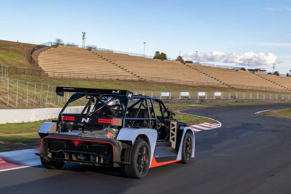 La imagen muestra un coche de carreras de alto rendimiento, específicamente el Hyundai RN24, en una pista. El coche tiene un diseño distintivo con una jaula antivuelco y características aerodinámicas, incluyendo un gran alerón trasero. El coche está marcado con la letra "N" en el alerón trasero y en el panel trasero. La pista tiene una curva y hay gradas vacías en el fondo, indicando un lugar de carreras. La escena se desarrolla durante el día con cielos despejados. Esta imagen es interesante porque captura un vehículo de alto rendimiento en un entorno de carreras, destacando los aspectos de ingeniería y diseño de los coches de carreras.