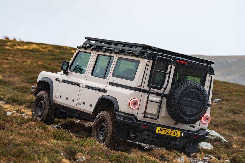 La imagen muestra un INEOS Grenadier, un vehículo todoterreno robusto, conduciendo sobre un terreno rocoso y cubierto de hierba. El vehículo es blanco con detalles en negro y cuenta con un portaequipajes en el techo, una rueda de repuesto montada en la parte trasera y una escalera en la parte posterior. La matrícula dice "HF72 FXU". Esta imagen destaca las capacidades todoterreno del vehículo y su diseño resistente, lo que lo hace relevante para aquellos interesados en aventuras fuera de carretera o vehículos duraderos. Además, el Grenadier está equipado con asientos RECARO, conocidos por su comodidad y soporte en condiciones extremas.