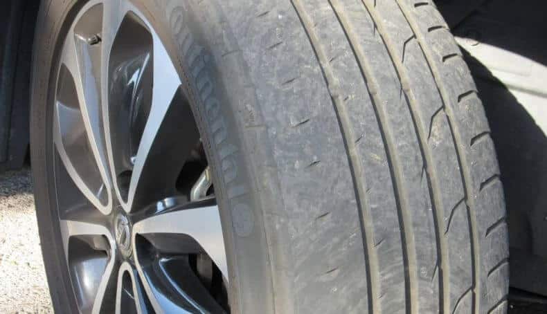 Primer plano fotorealista de un neumático extremadamente desgastado en formato 16:9. La sección central está completamente calva con los cables de acero visibles, la profundidad de la banda de rodadura está muy por debajo del límite legal de 1.6 mm, con los indicadores de desgaste completamente borrados. No hay secciones de neumático nuevo visibles. El patrón de la banda de rodadura restante es casi inexistente, mostrando una superficie vidriosa y pulida por el desgaste extremo. Se observan múltiples grietas, cortes y escombros incrustados en el caucho restante. Iluminación profesional de estudio para resaltar la textura peligrosamente lisa de la superficie. Un medidor de profundidad muestra una medida crítica de 0.5 mm. Fotografía tomada con lente macro, enfocando en las áreas más desgastadas.