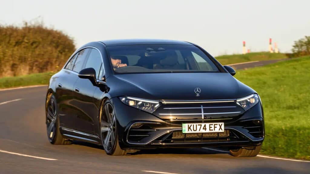 Un Mercedes-Benz EQS negro conduciendo por una carretera. El coche es un vehículo eléctrico moderno y elegante con una parrilla frontal distintiva y faros LED. La matrícula dice 'KU74 EFX'. El coche está en una carretera sinuosa con césped verde a ambos lados, lo que sugiere que está siendo conducido en un área rural o suburbana. Esta imagen es interesante y relevante ya que muestra un vehículo eléctrico de lujo, destacando los avances en tecnología automotriz y la transición hacia el transporte sostenible.