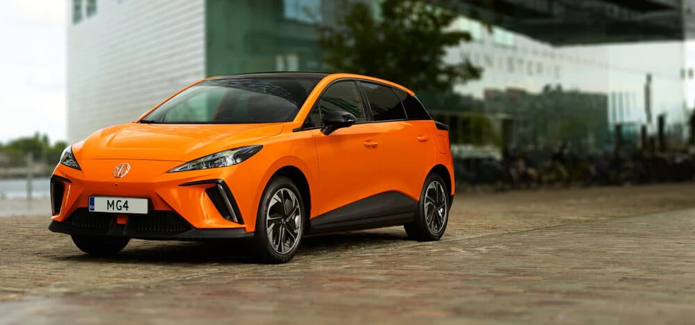 Imagen de un MG4 naranja estacionado sobre una superficie de adoquines. El coche tiene un diseño moderno y elegante con líneas afiladas y faros distintivos. Al fondo, se puede ver un edificio moderno con vidrio reflectante y algo de vegetación. La matrícula del coche dice 'MG4'. Esta imagen es interesante y relevante ya que muestra el diseño y la estética del modelo MG4, destacando su apariencia contemporánea y estilizada.