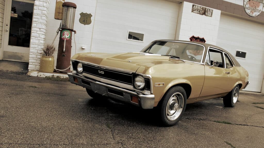 Imagen de un coche clásico, específicamente un Chevrolet Nova SS, estacionado frente a una gasolinera de estilo antiguo. El coche es de color beige con detalles cromados y tiene la insignia 'SS' en la parrilla delantera, indicando que es un modelo Super Sport. La gasolinera cuenta con una bomba de gasolina vintage y varios letreros retro, incluyendo uno de la carretera 'Georgia US 41' y otro de 'Gasoline'. El ambiente evoca una sensación de nostalgia, capturando la esencia de una era pasada en la historia automotriz y la cultura americana.