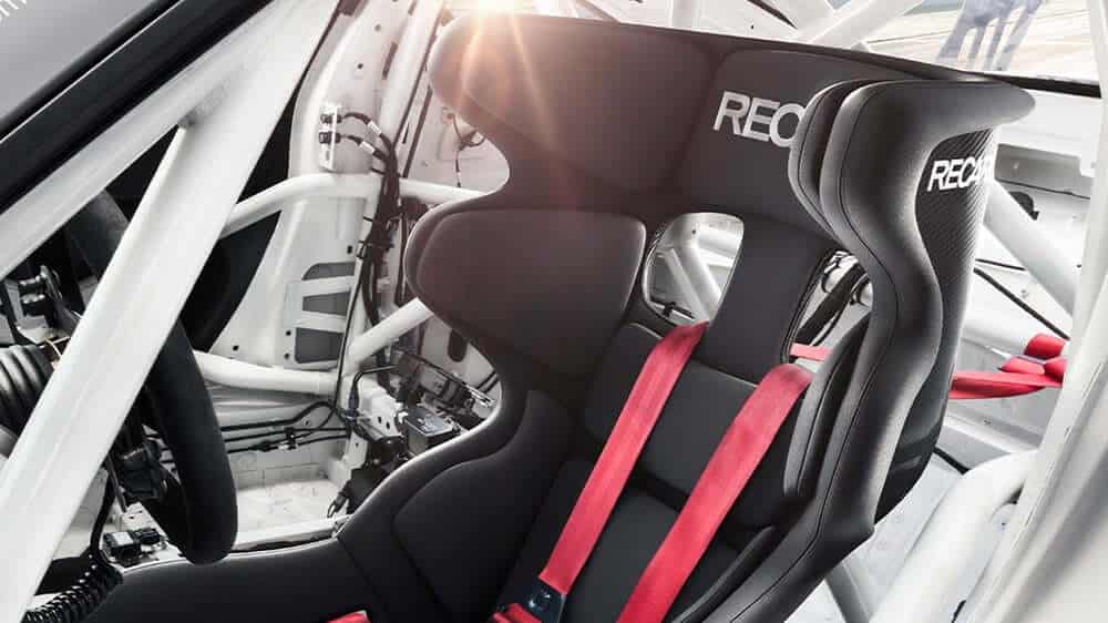 La imagen muestra el interior de un coche de carreras, centrado en un asiento de carreras RECARO. El asiento está diseñado para conducción de alto rendimiento, con un respaldo alto y protección lateral para la cabeza, y está equipado con arneses de seguridad rojos. El interior del coche también incluye una jaula antivuelco para mayor integridad estructural y seguridad durante las carreras. La marca RECARO es visible en el reposacabezas del asiento. Este conjunto es interesante y relevante para aquellos involucrados en el automovilismo o entusiastas del automóvil que aprecian la importancia de la seguridad y el rendimiento en entornos de carreras.