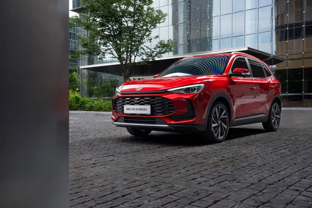 La imagen muestra un MG ZS Hybrid+ rojo estacionado sobre una superficie de adoquines frente a un edificio moderno de vidrio. El vehículo se destaca por su diseño elegante y su tecnología híbrida. El entorno sugiere un ambiente urbano, y las características modernas y el sistema híbrido ecológico del coche lo hacen relevante para discusiones sobre tendencias automotrices contemporáneas y transporte sostenible.