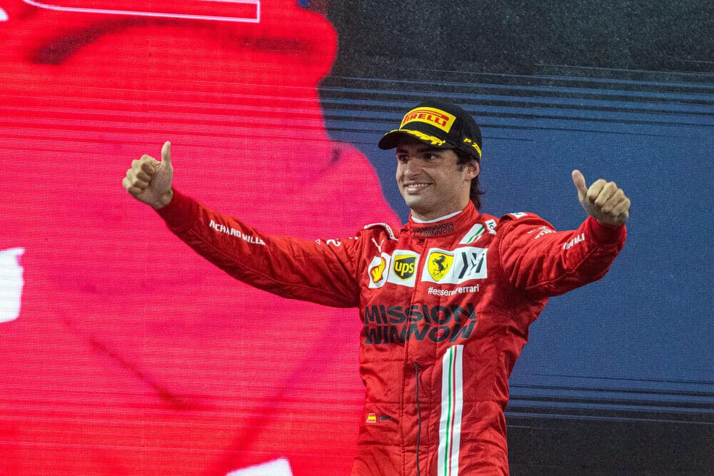Gracias por la aclaración. La imagen muestra a Carlos Sainz, piloto de Fórmula 1, con un traje de carreras rojo que lleva varios logotipos de patrocinadores, incluyendo Shell, UPS y Ferrari. Carlos Sainz está haciendo un gesto de doble pulgar hacia arriba y lleva una gorra negra con el logotipo amarillo de Pirelli. El fondo es predominantemente rojo con algunos elementos en negro y blanco. Esta imagen es interesante y relevante ya que representa un momento de celebración o victoria en un evento de automovilismo, probablemente relacionado con la Fórmula 1, dado el branding de Ferrari.