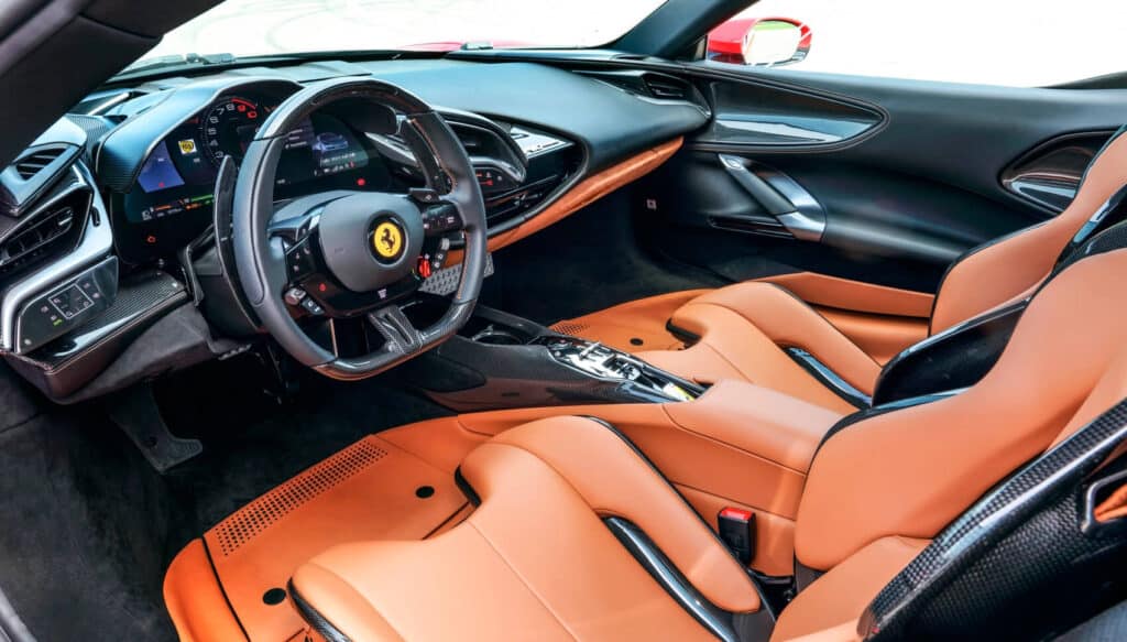 La imagen muestra el interior de un Ferrari SF90 Stradale. El diseño interior es lujoso y deportivo, con asientos de cuero naranja brillante y detalles en fibra de carbono en la consola central. El volante lleva el logotipo de Ferrari y varios controles, incluyendo las levas de cambio. El tablero de instrumentos es digital y muestra información esencial del vehículo. La combinación de materiales de alta calidad y tecnología avanzada hace que este interior sea tanto visualmente atractivo como funcional para la conducción de alto rendimiento.