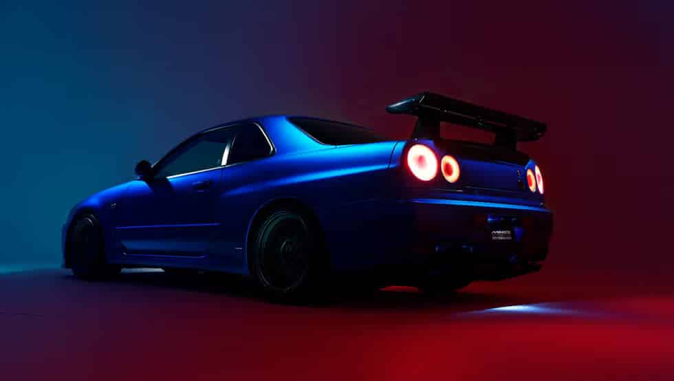 Gracias por la corrección. La imagen muestra un Nissan Skyline GT-R R34 modificado por Built by Legends. El coche deportivo azul tiene un prominente alerón trasero y está iluminado por una combinación de luces azules y rojas. La vista desde el lado trasero izquierdo destaca su diseño aerodinámico y sus distintivas luces traseras redondas. La iluminación y el ángulo enfatizan la forma aerodinámica y las características orientadas al rendimiento del coche, haciéndolo visualmente llamativo y atractivo para los entusiastas del automovilismo.