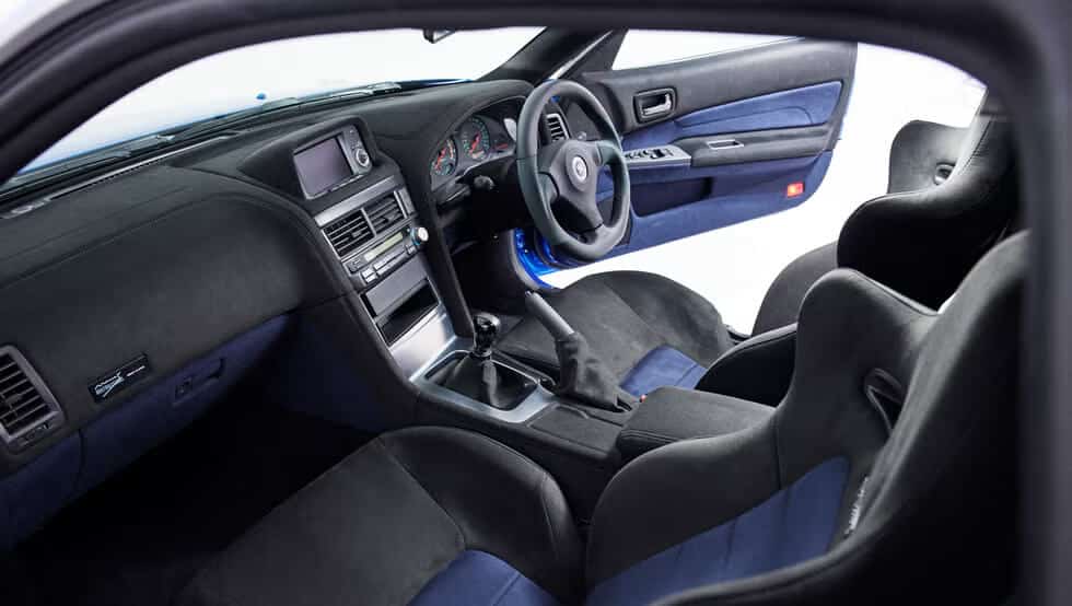 La imagen muestra el interior de un Nissan Skyline GT-R R34. El coche tiene una configuración de volante a la derecha, lo que indica que está diseñado para países donde se conduce por el lado izquierdo de la carretera. El interior presenta una tapicería en negro y azul, una palanca de cambios manual y un tablero moderno con una pantalla digital y varios controles. Los asientos parecen ser de estilo deportivo, lo que sugiere que el coche podría ser un modelo de alto rendimiento. Esta imagen es interesante porque destaca el diseño y las características del interior de un vehículo icónico y de alto rendimiento, muy apreciado por los entusiastas del automovilismo.
