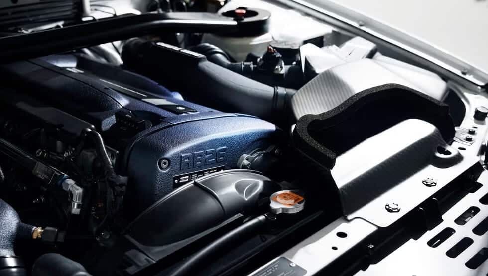 La imagen muestra el compartimento del motor de un coche, destacando específicamente el motor RB26. Este motor es bien conocido por ser producido por Nissan y está asociado frecuentemente con vehículos de alto rendimiento como el Nissan Skyline GT-R. El RB26 es notable por su configuración de seis cilindros en línea con doble turbo, lo que lo convierte en una opción popular entre los entusiastas de los coches y los tuners debido a su potencial para producir una potencia significativa. La imagen captura varios componentes del motor, incluyendo el colector de admisión, los turbocompresores y otras partes relacionadas, mostrando la naturaleza intrincada y de alto rendimiento del motor RB26.
