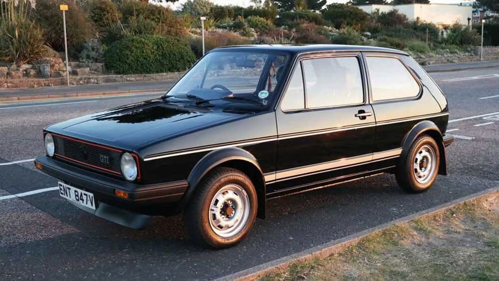 La imagen muestra un Volkswagen Golf GTI Mk1 negro estacionado al lado de una carretera. El coche tiene un diseño clásico con faros redondos, una franja roja alrededor de la parrilla delantera y una distintiva insignia "GTI". La matrícula dice "ENT 847V".