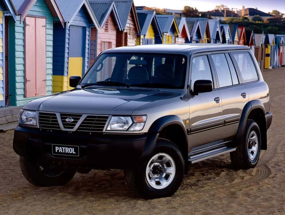 Un Nissan Patrol Y61 de 1997 está estacionado en una playa de arena frente a una fila de coloridas cabañas de playa. El vehículo es un SUV grande de cuatro puertas con un diseño robusto y resistente, indicativo de sus capacidades todoterreno. Las cabañas de playa en el fondo están pintadas en varios colores brillantes, añadiendo un elemento vibrante y pintoresco a la escena. Esta imagen destaca la idoneidad del Nissan Patrol Y61 para entornos al aire libre y aventureros, enfatizando su durabilidad y versatilidad.