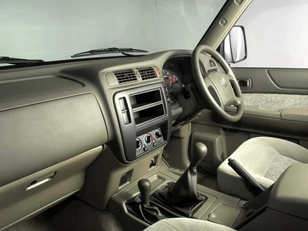 "El interior de un Nissan Patrol Y61 de 1997 visto desde el lado del pasajero delantero. Se muestra el tablero de instrumentos, el volante y los asientos delanteros. El tablero tiene un diseño simple con una ranura vacía para un sistema de estéreo, controles de aire acondicionado y varios botones y diales. El volante tiene el logotipo de Nissan en el centro. La palanca de cambios y el freno de mano son visibles entre los asientos delanteros. El interior es principalmente gris con asientos de tela.
