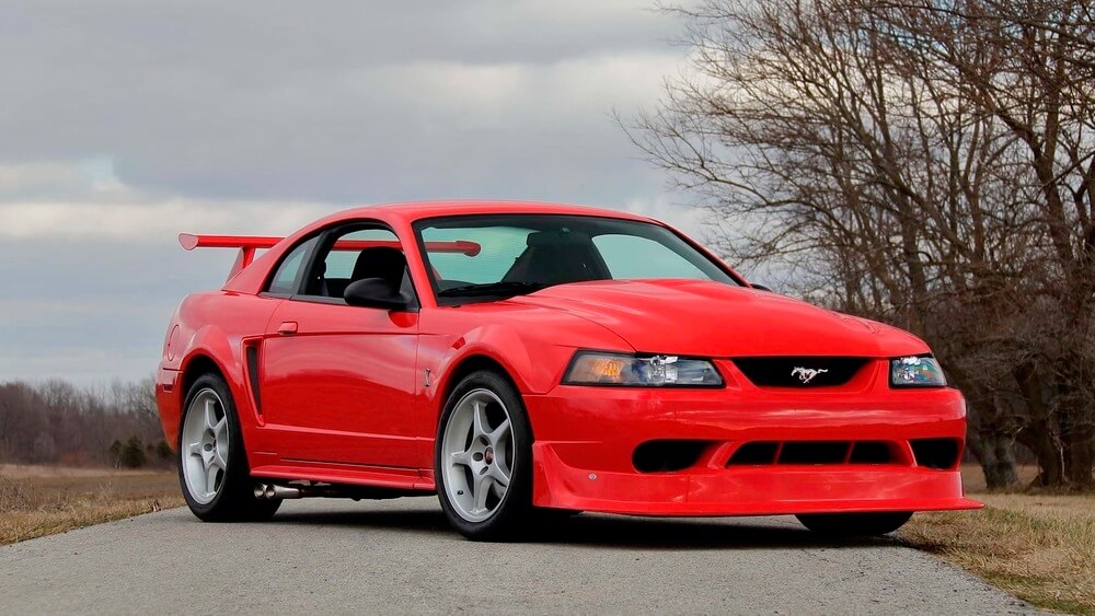 Imagen de un Ford Mustang SVT Cobra R del año 2000 de color rojo brillante, estacionado en una carretera con un fondo natural de árboles sin hojas y un cielo nublado. El coche parece estar en excelente estado, con un exterior brillante y un diseño deportivo que incluye un parachoques delantero prominente, faldones laterales y un gran alerón trasero. El emblema del Mustang es visible en la parrilla frontal.