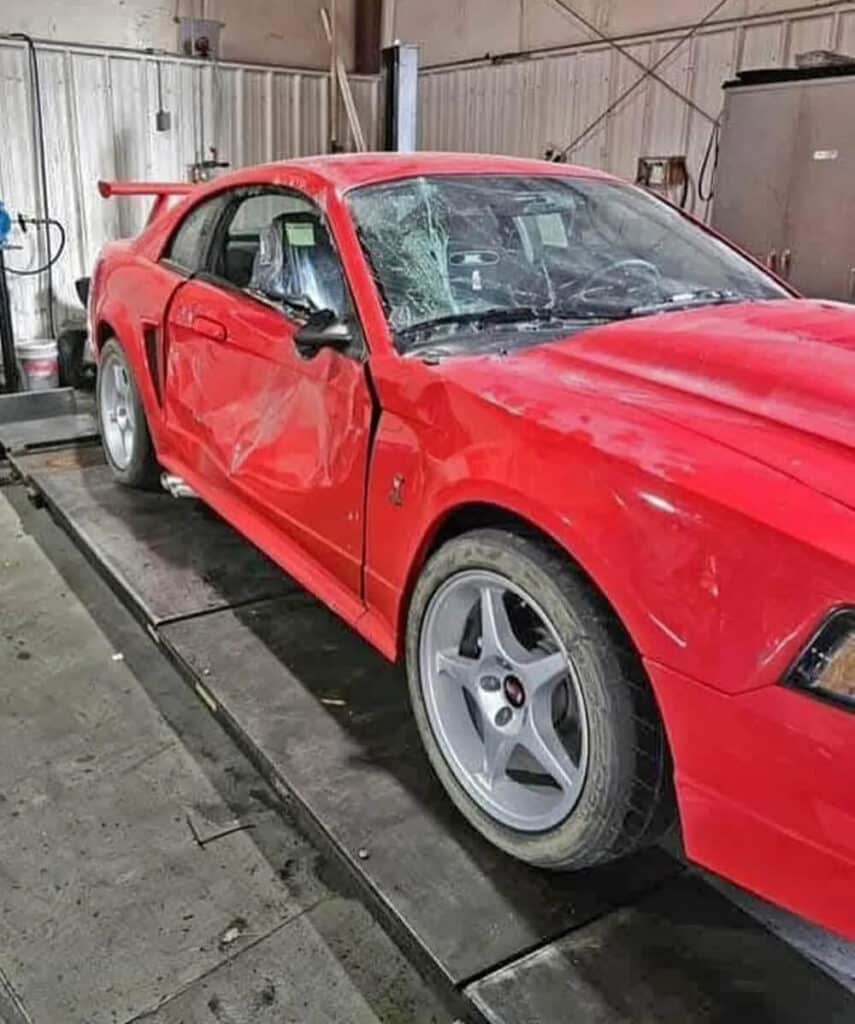 Imagen de un Ford Mustang SVT del año 2000 de color rojo, con daños visibles. El coche tiene una gran abolladura en la puerta del lado del conductor y el parabrisas está roto. El vehículo está dentro de un garaje o taller, probablemente para reparaciones. Los daños sugieren que el coche estuvo involucrado en un accidente.