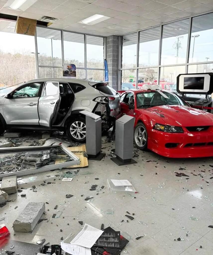 Esquema de una colisión en el concesionario Tri-City Ford. El diagrama muestra la disposición del concesionario, incluyendo el "Showroom Floor" y las áreas del "Tri-City Ford Dealership". El Ford Mustang SVT está estacionado y no se mueve. El Hyundai Tucson ha atravesado el escaparate del concesionario, causando daños significativos en la parte trasera de la Tucson y en el lateral derecho del Mustang. No hay personas presentes en la escena.