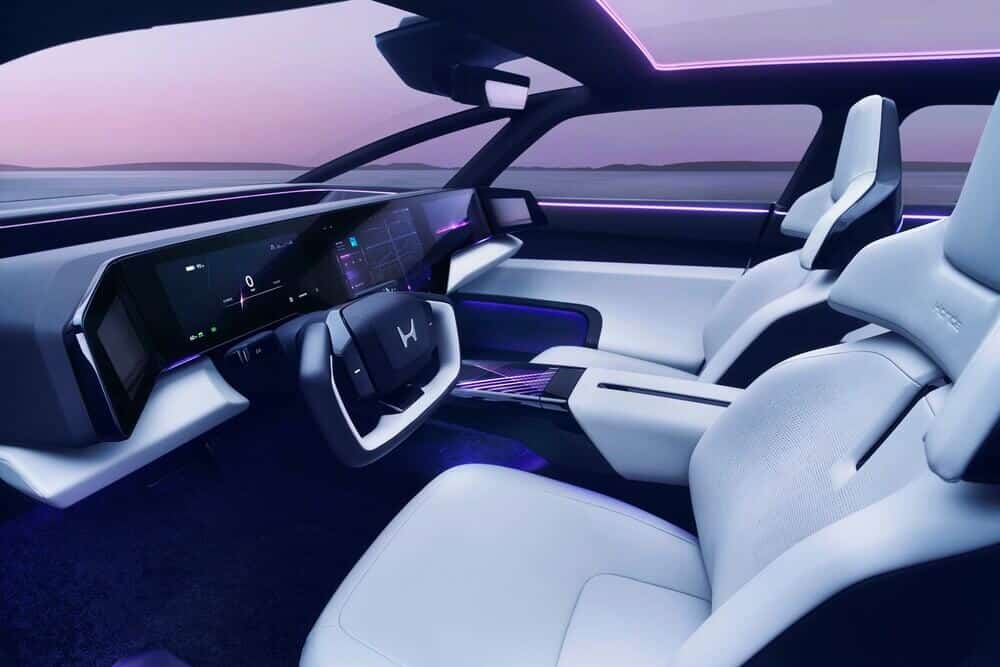 Imagen del interior de un automóvil Honda 0 Saloon. El interior presenta un diseño moderno y elegante con tecnología avanzada. El tablero está dominado por una gran pantalla digital continua que abarca todo el ancho del coche, mostrando información como navegación y estado del vehículo. El volante tiene un diseño único con un logotipo central. Los asientos son ergonómicos y elegantes, con un esquema de colores blanco y negro. La iluminación ambiental es de color púrpura, lo que añade un toque de alta tecnología y lujo al interior.