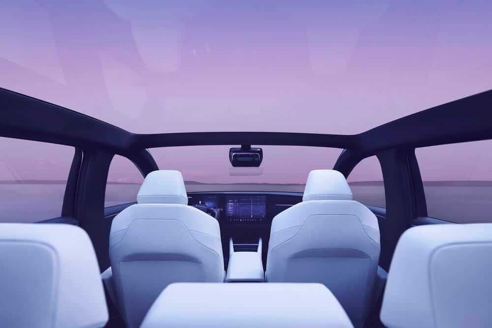 "Imagen del interior de un automóvil Honda 0 Saloon, vista desde el asiento trasero hacia adelante. El coche cuenta con un techo panorámico de vidrio, proporcionando una vista clara del cielo, que está teñido con un degradado de tonos púrpura. Los asientos delanteros son blancos y tienen un diseño elegante y minimalista. El tablero está equipado con una gran pantalla táctil central, que parece mostrar interfaces de navegación o control. El diseño general del interior del coche es futurista y lujoso, enfatizando la tecnología avanzada y la comodidad.