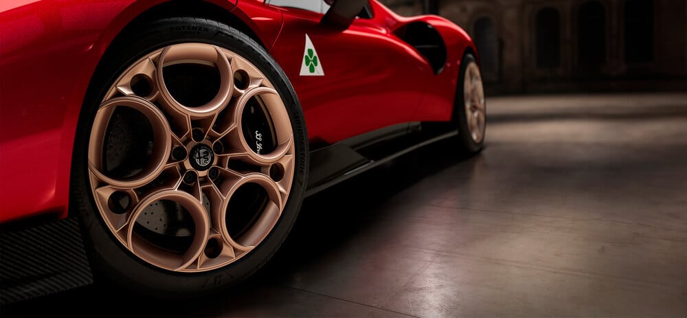 Imagen de un primer plano de la rueda delantera de un auto deportivo rojo Alfa Romeo 33 Stradale. La rueda tiene un diseño distintivo con múltiples recortes circulares y un acabado en bronce. El auto tiene un emblema de trébol verde en el lateral, indicando que es un modelo de Alfa Romeo. El entorno parece ser interior, con un suelo pulido y un fondo con poca luz, destacando la apariencia elegante y estilizada del auto.
