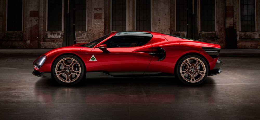 Imagen de un auto deportivo rojo Alfa Romeo 33 Stradale con un diseño moderno y aerodinámico. El auto está posicionado en un entorno industrial con poca luz, lo que resalta su acabado brillante y detalles intrincados. El auto presenta un logotipo triangular distintivo en el lateral. Las ruedas tienen un diseño único y elegante, y la apariencia general del auto sugiere alto rendimiento y lujo.