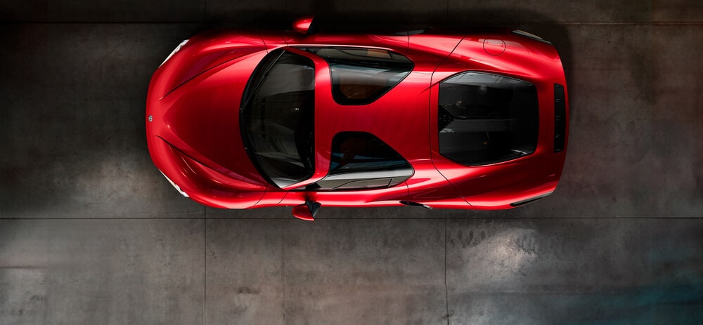 Imagen de vista aérea de un auto deportivo rojo Alfa Romeo 33 Stradale. El auto tiene un diseño aerodinámico con curvas suaves y un acabado brillante. El techo presenta grandes paneles de vidrio, probablemente parte de un techo panorámico, y la forma aerodinámica del auto se enfatiza con sus líneas y contornos afilados. El entorno parece ser un garaje o sala de exposición interior con un suelo de concreto.