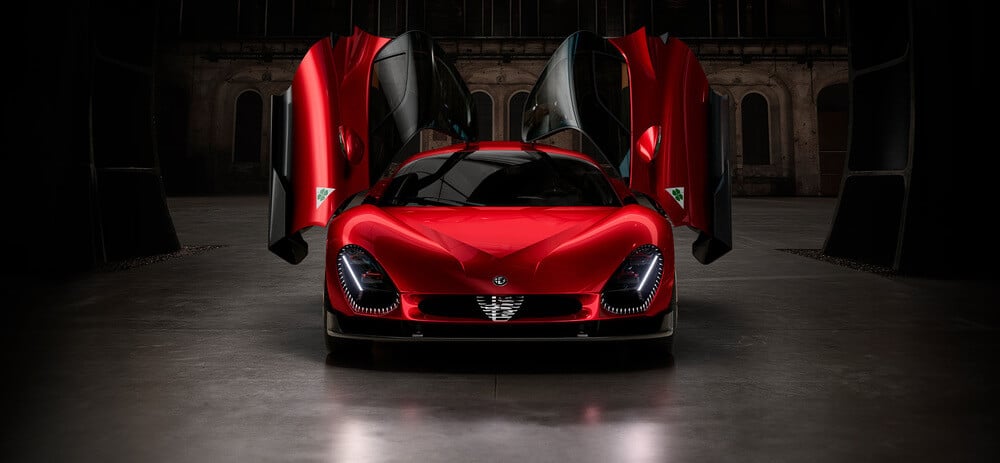 Imagen de un auto deportivo rojo Alfa Romeo 33 Stradale con puertas de ala de mariposa abiertas, mostrando un diseño aerodinámico y elegante. El auto está en un entorno industrial con poca luz, lo que resalta su acabado brillante y estética moderna. El frente del auto presenta faros distintivos y una parrilla prominente con un logotipo en el centro.