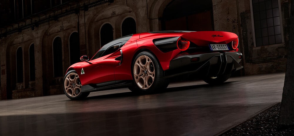 Imagen de un auto deportivo rojo Alfa Romeo 33 Stradale con características de diseño distintivas, incluyendo una carrocería aerodinámica, un gran difusor trasero y luces traseras únicas. El auto está estacionado frente a un edificio industrial antiguo con ventanas arqueadas y una fachada desgastada. La placa del auto dice 'AR 0333T'. El diseño del auto y el entorno crean un contraste llamativo entre la ingeniería automotriz moderna y la arquitectura histórica.